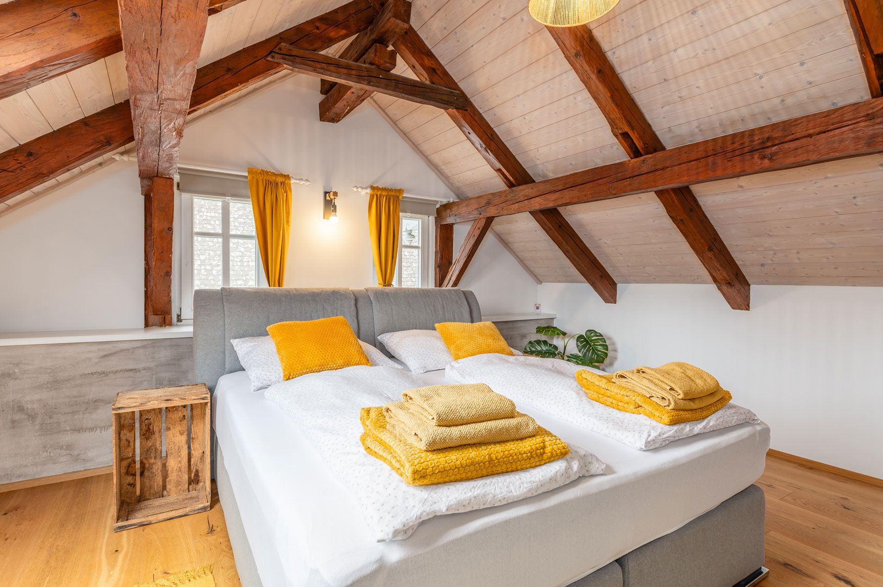 ferienwohnung-hoamat-fuers-herz-doppelbett-dachgeschoss-groebming-ennstal-Die-große-Hoamat Dachgeschoss-Schlafzimmer in Ferienwohnung mit Doppelbett, grauem Kopfteil, gelben Kissen und Handtüchern, Holzbalken und zwei Fenstern mit gelben Vorhängen